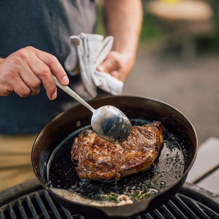 Person spoons pan juices over a ribeye steak searing in a Field Cast Iron Skillet on an outdoor grill