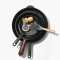 Three cast iron skillets showing a slotted turner, wooden brush, seasoning oil, mesh scrubber, and leather handle cover inside. thumbnail